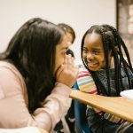 Two girls whisper and smile in a classroom, fostering friendship and joy.